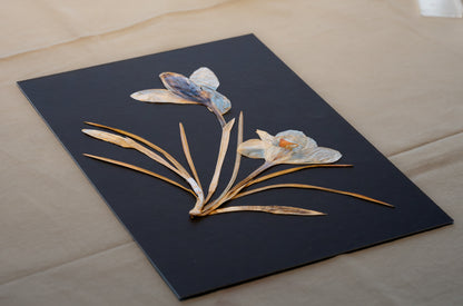 Process of framed arwork, wall decor to gift for loved ones and home decor for loved homes. A square black wall art panel featuring an intricate floral arrangement made from natural materials, including dried iris petals, leaves, and a small butterfly embellishment, displayed on a neutral fabric surface.