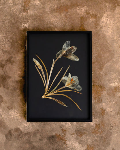 A framed botanical art print featuring pressed flowers with metallic gold and gray-green petals on a black background, set against a textured brown wall. Gift for loved ones, home decor for your loved home. 