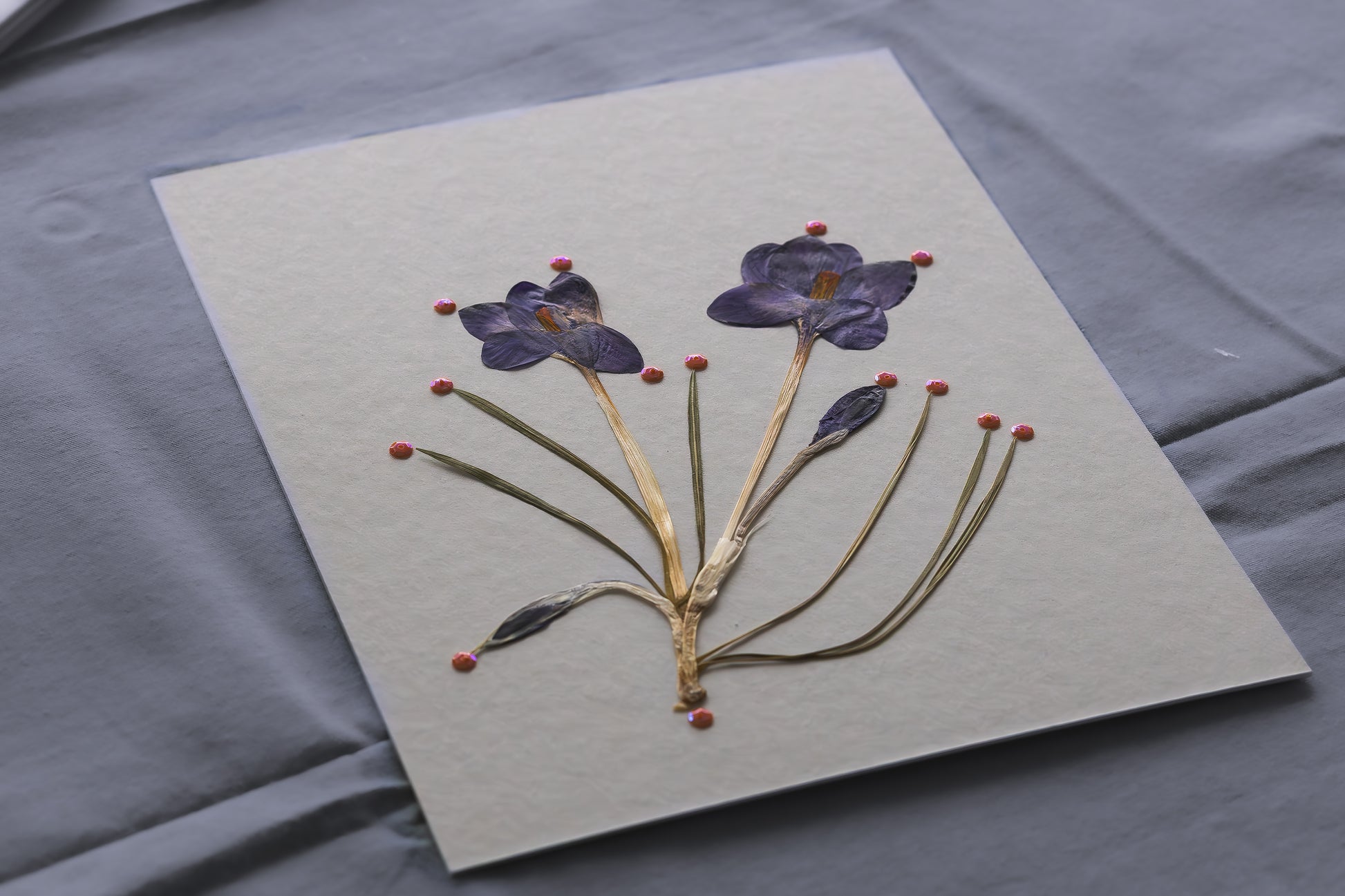 A square handmade paper card featuring pressed purple flowers and green stems, displayed on a textured gray fabric background. Process of framed botanical artwork as a gift or home decor