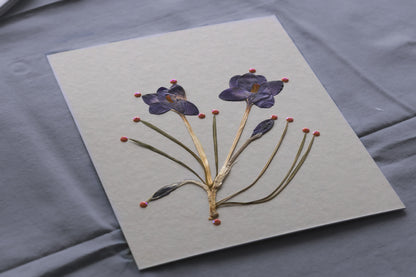 A square handmade paper card featuring pressed purple flowers and green stems, displayed on a textured gray fabric background. Process of framed botanical artwork as a gift or home decor