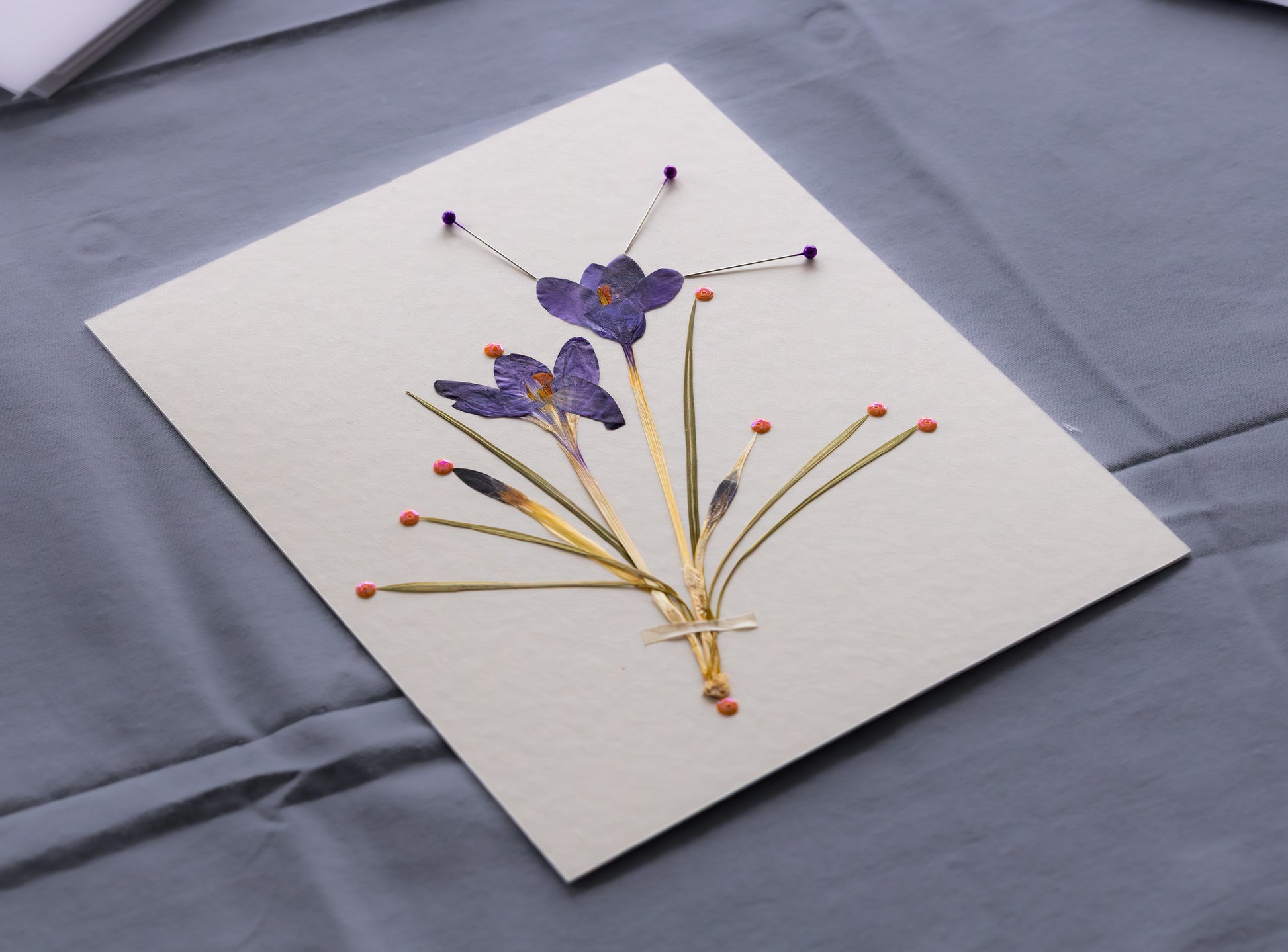 A square white card featuring pressed purple flowers and green stems arranged on a gray fabric surface.